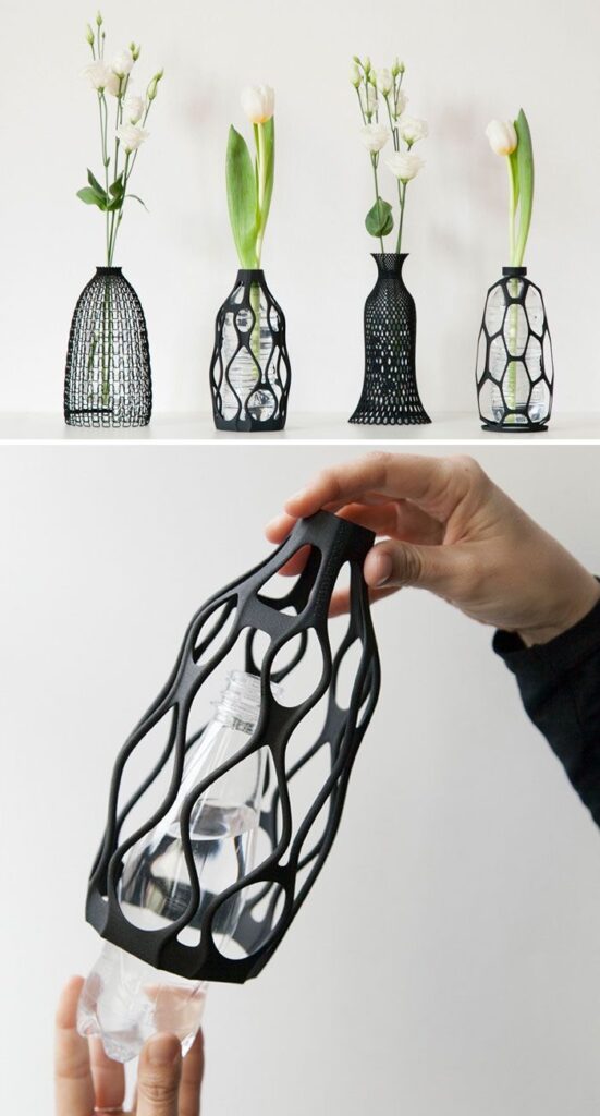 A hand holds a black, lattice-work 3d printed vase containing a white tulip, with more vases and flowers blurred in the background.