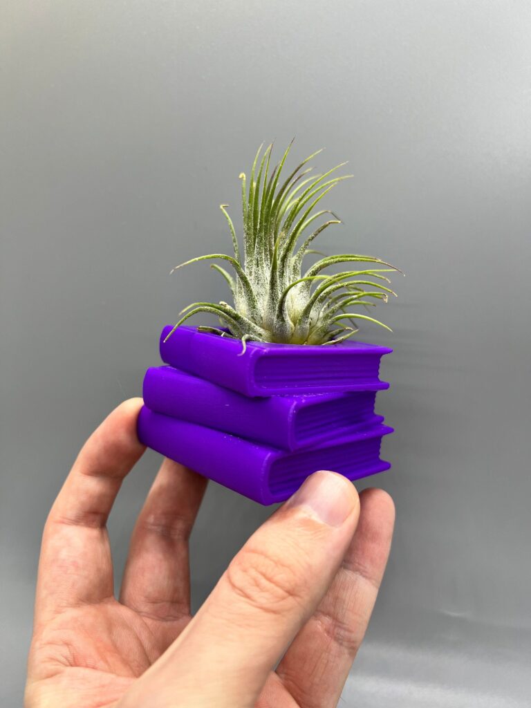 A hand holding a small purple 3d-printed book-shaped planter containing an air plant, against a gray background.