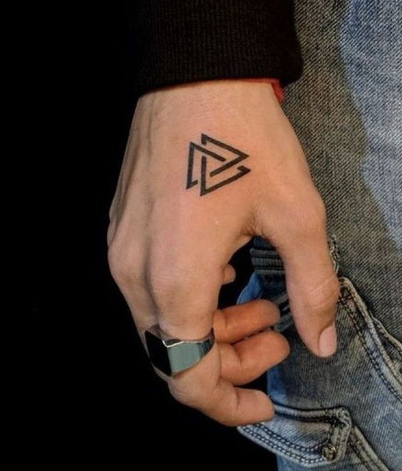 A close-up of a person's hand with a geometric arrow tattoo on the back, resting against denim jeans. they are wearing a silver ring on their thumb.