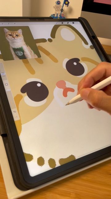 A hand using a stylus to draw a cartoon cat on a digital tablet, with a photo of a real cat wearing a starbucks apron visible in the corner.