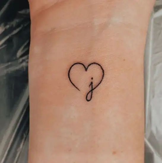 A small, black tattoo of a heart with a lowercase letter 'j' inside, inked on a person's wrist.