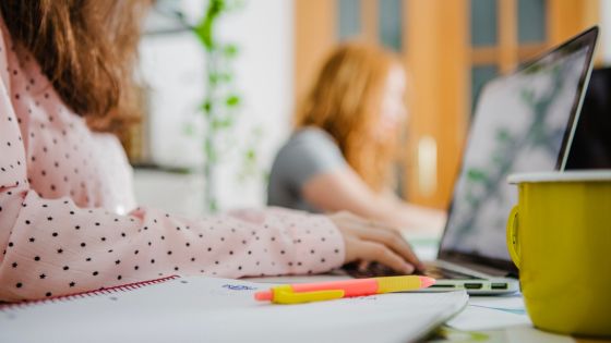 A woman in a polka-dot shirt working on a laptop with another woman in the background also using a laptop. a coffee mug and stationery are on the table.