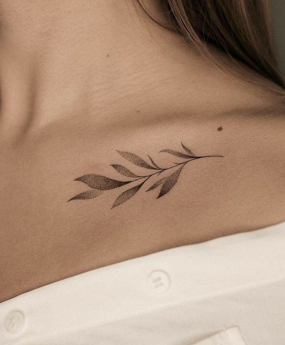 Close-up of a woman's collarbone with a delicate leaf tattoo on her skin, partially obscured by the collar of a white shirt.