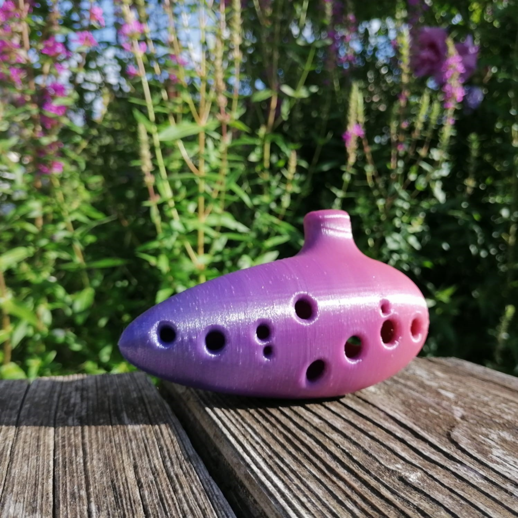A purple ceramic ocarina with multiple holes placed on a wooden surface, surrounded by colorful flowers and greenery in the background.