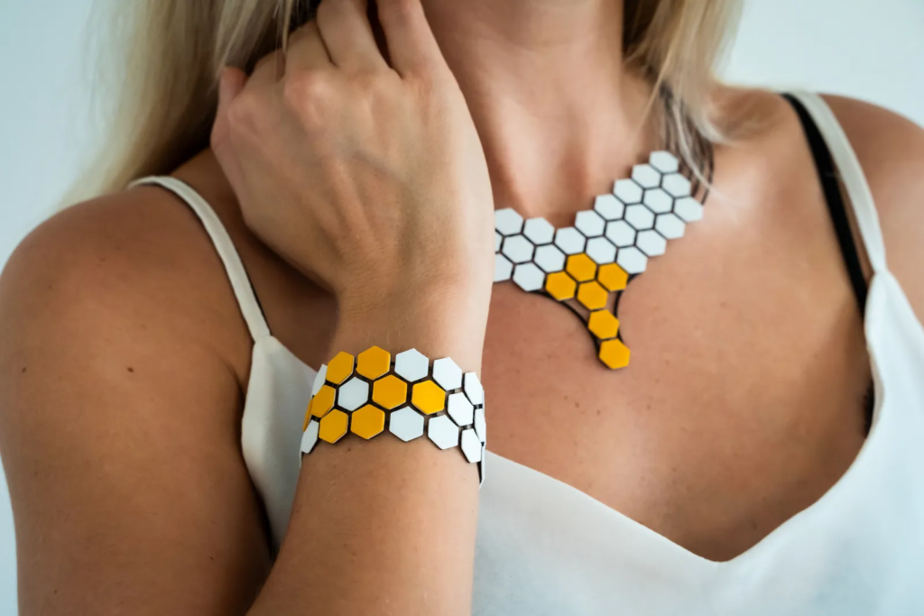 Close-up of a woman with blonde hair wearing a geometric, hexagonal-patterned necklace and bracelet set, featuring predominantly white and a few yellow tiles.