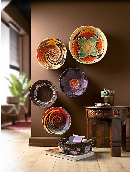 Five woven baskets with colorful patterns are mounted on a brown wall. A wooden table with books and a plant sits nearby on a light wood floor, next to a basket with additional items.