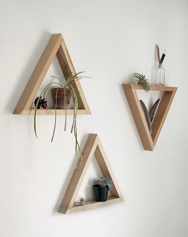 Three triangular wooden wall shelves holding small potted plants, a feather in a glass jar, and a pinecone, mounted on a white wall.