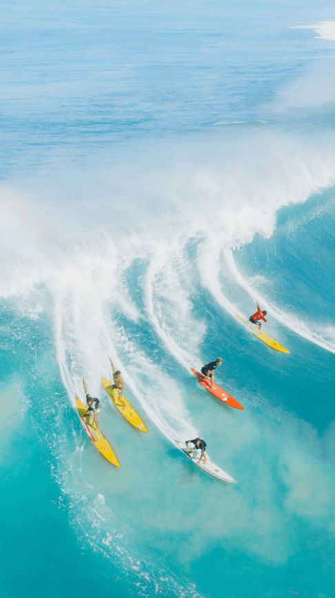 Five surfers ride large waves in the ocean, each on their own brightly colored surfboard, creating trails of white spray behind them. The water is blue and the sky is clear.