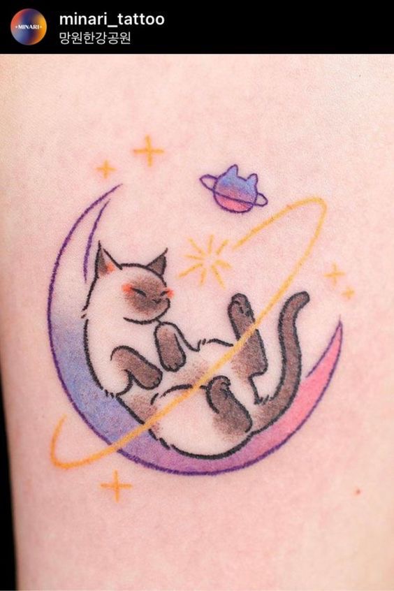 A tattoo of a sleeping cat floating within a crescent moon, encircled by orange rings, next to a small purple planet.