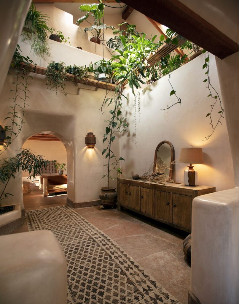 A well-lit hallway in a rustic home with white stucco walls, a patterned rug, potted plants, wooden furniture, and a lamp. An archway leads to another room with more plants and natural decor.