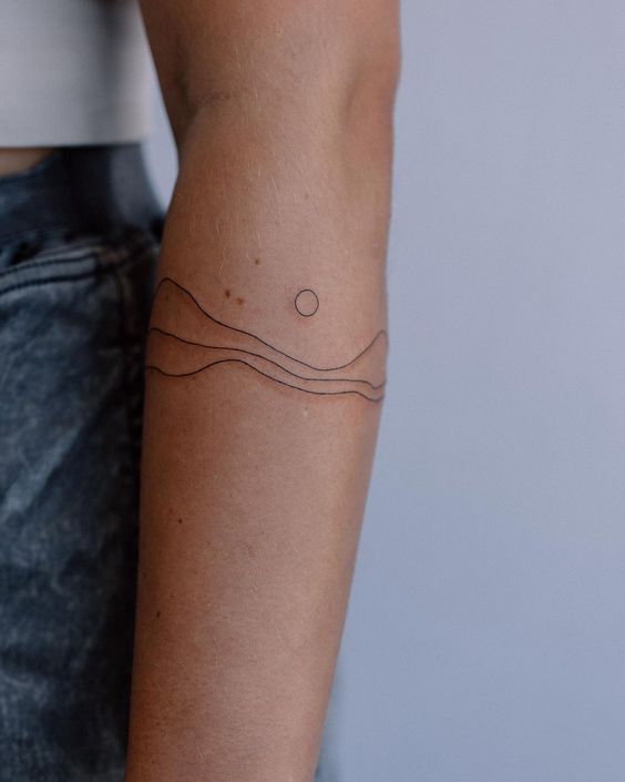 Close-up of a person's arm with a minimalist tattoo consisting of three wavy lines and a small circle above them. The person is wearing a cropped top and denim shorts.