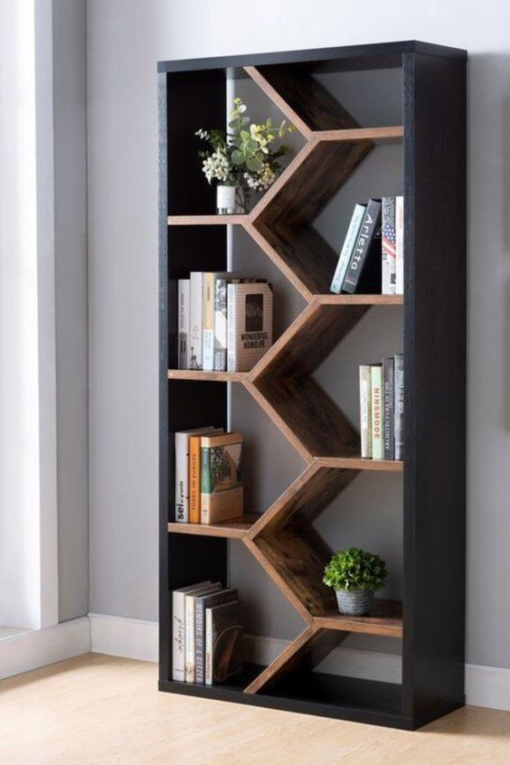 A modern bookshelf with a geometric zigzag design, containing plants, books, and decorative items, set against a white wall on a hardwood floor.