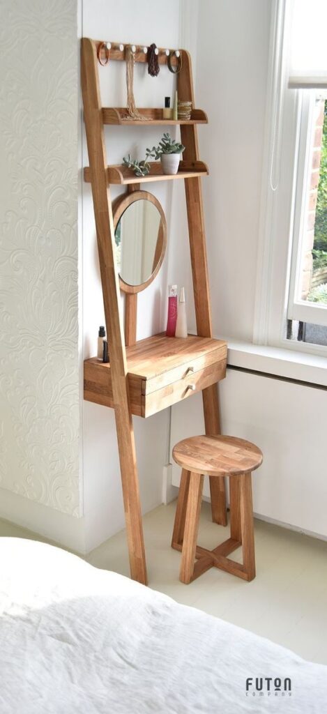 A wooden vanity with a mirror and drawer, adorned with accessories, and a matching stool is set against a white wall near a window.