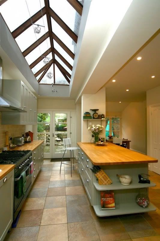 A modern kitchen with a long central island, skylight ceiling, and tile floor. Shelves on the island hold books and various items. The kitchen leads to a door opening to an outdoor area.