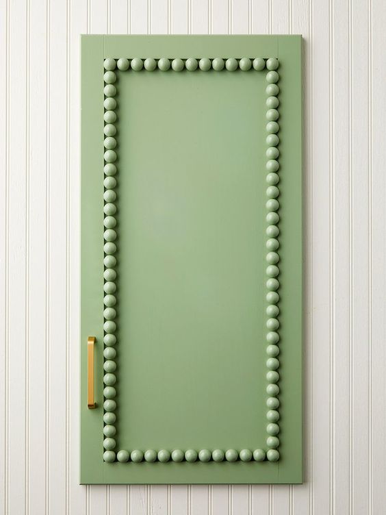 A green cabinet door with a rectangular border of round knobs and a brass handle is mounted on a vertically-striped white wall.