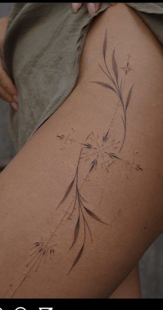 Close-up of a leg with a delicate floral tattoo, featuring thin branches and leaves with star-like elements, displayed against a background of textured skin.