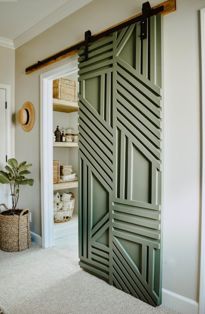 Green sliding barn door with a geometric pattern partially closed, revealing a pantry with woven storage baskets and neatly arranged items. A potted plant is placed nearby.