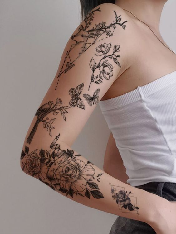 A person with a detailed black ink tattoo sleeve featuring flowers, butterflies, a bird, and geometric shapes on their right arm, wearing a white sleeveless top.