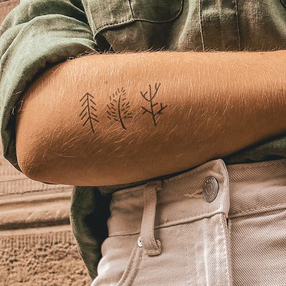 An arm with four minimalistic tree tattoos crossed over a torso wearing a green shirt and white pants.