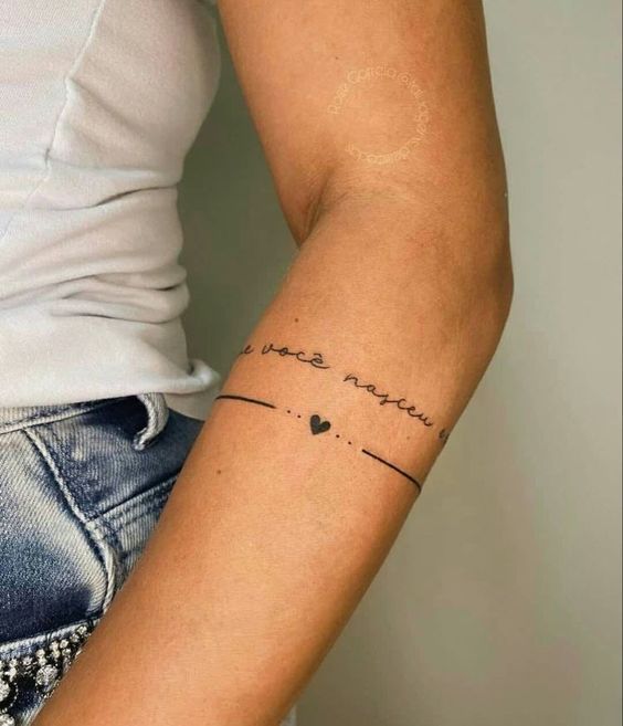 A person wearing a white shirt displays a tattoo on their arm featuring a curved line with small dots and a heart, accompanied by cursive text.