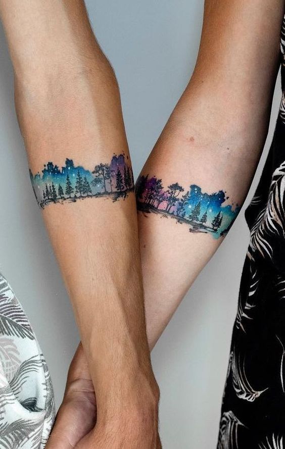Two forearms with matching tree-line tattoos in watercolor style, featuring shades of blue, purple, and green. Arms are crossed, showcasing the tattoos side by side.
