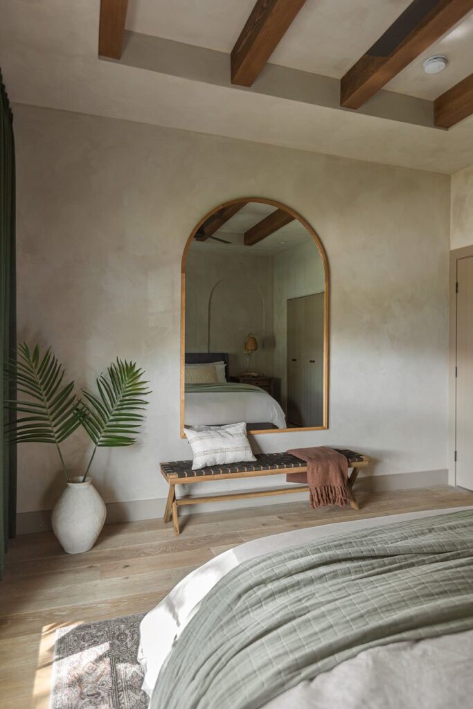 A bedroom features a large arched mirror above a bench with pillows and a throw blanket. A potted plant is in the corner, and the bed has green and white bedding. Exposed wooden beams are on the ceiling.