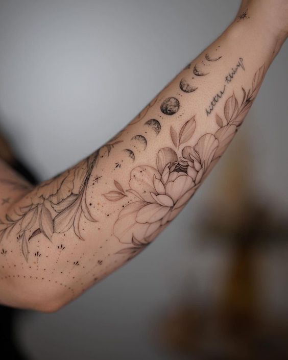 A forearm tattoo featuring phases of the moon, floral designs, and the handwritten text "moon thing" on the inner arm.