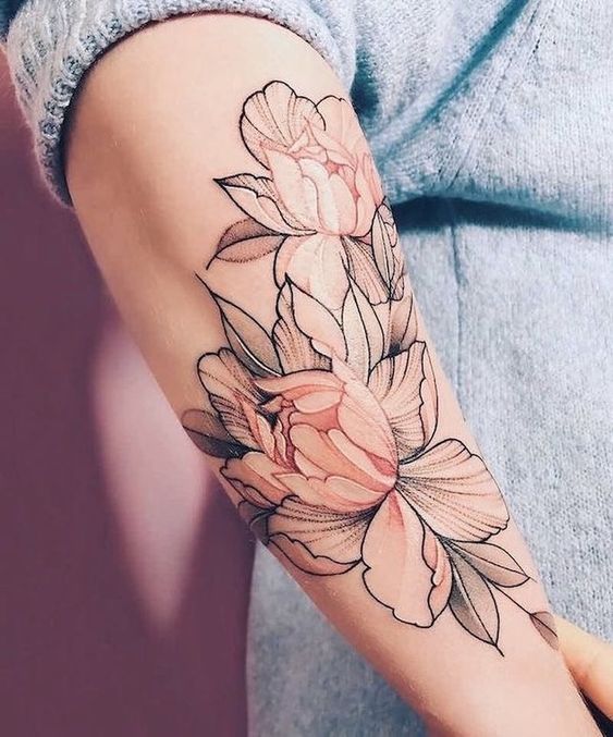 Tattoo of large, pink flowers with green leaves covers a person's forearm. The person is wearing a light blue, long-sleeved shirt.