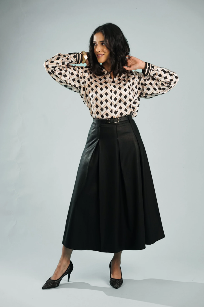 Woman standing, posing in a patterned blouse, black leather skirt, and heels, with her hands near her head, smiling, against a grey background.