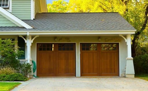 A residential house with a double garage door made of brown wood, surrounded by greenery and a paved driveway in a suburban setting.