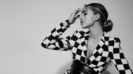 Woman poses in a black and white checkered blazer with one hand resting on her forehead against a plain background, black and white photo.