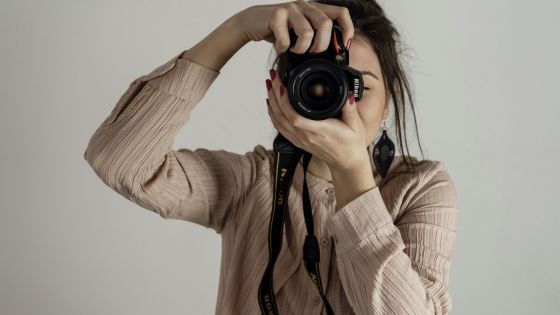 Person in a beige top holding a camera to their face, looking through the viewfinder, against a plain background.