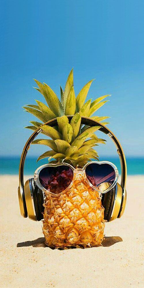 A pineapple wearing sunglasses and headphones is placed on a sandy beach with a clear blue sky in the background.