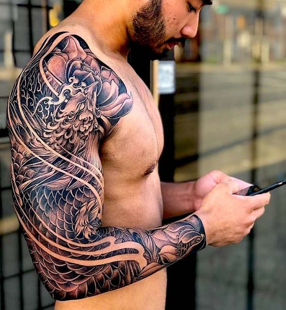 A shirtless man with a detailed dragon tattoo sleeve on his left arm is looking down at and using his phone.