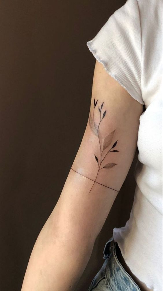 Upper arm with a minimalist tattoo of a plant with leaves, featuring shaded areas, on a person wearing a white short-sleeve shirt and blue jeans.