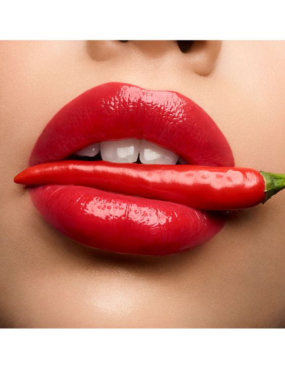 Close-up of glossy red lips holding a red chili pepper horizontally between them.