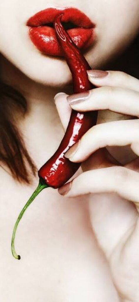 A close-up of a person holding a red chili pepper near their red-painted lips.
