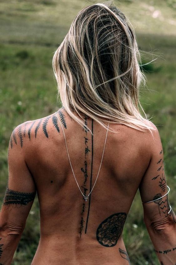 A back view of a person with long, blonde hair, multiple tattoos, including geometric and arrow designs, and a silver necklace standing outdoors.