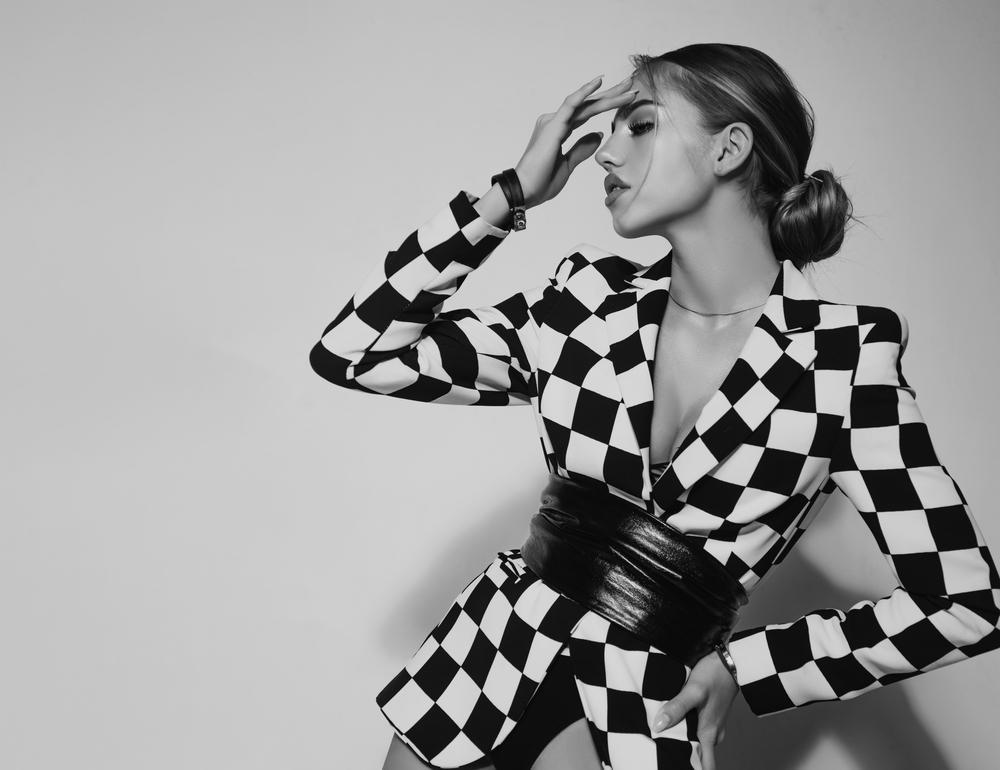 A woman in a black and white checkered blazer poses with one hand on her forehead and the other on her waist, looking to the left against a plain backdrop.
