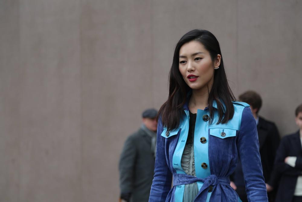 A woman with straight, shoulder-length hair wears a blue trench coat. She stands outdoors against a plain background with two people blurred in the background.