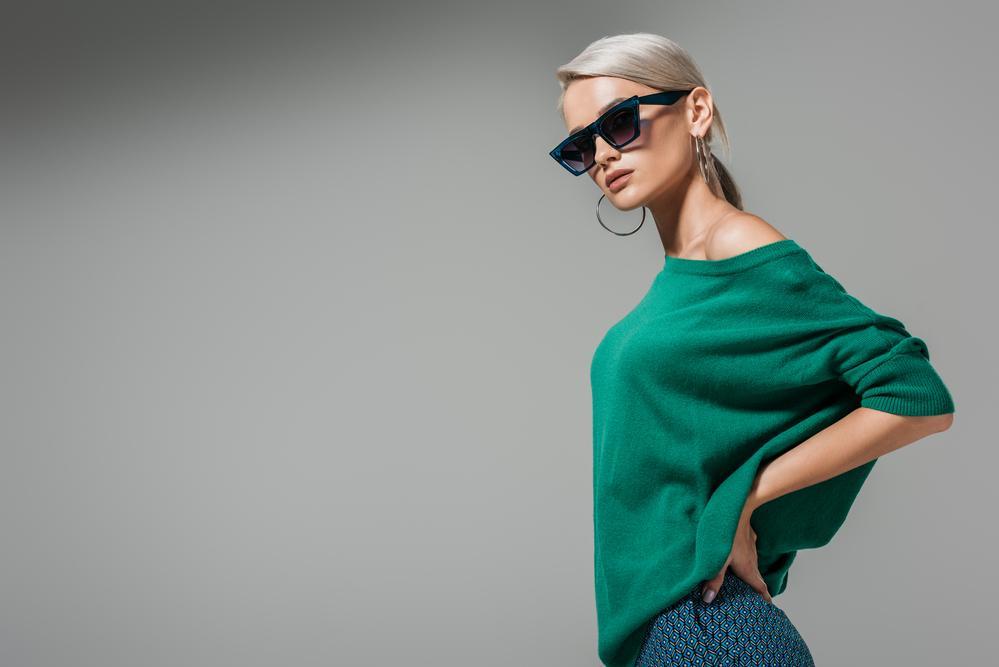 Woman wearing large sunglasses and an off-shoulder green sweater stands with one hand on her hip against a plain background.