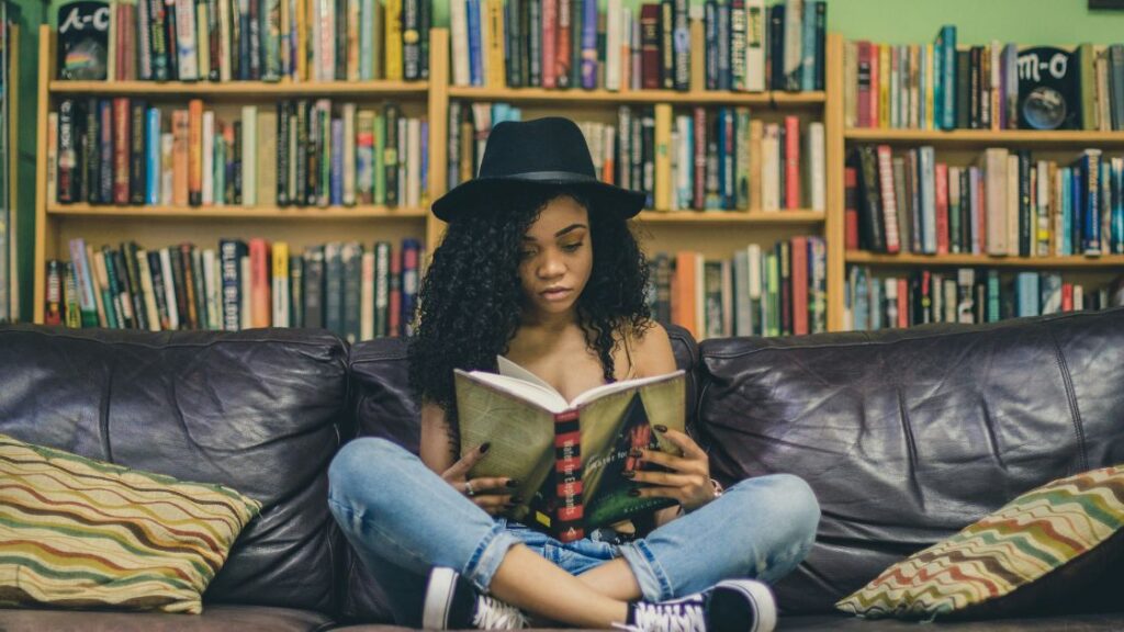 A person with long curly hair is sitting cross-legged on a brown couch, reading a book. They are wearing a black hat, blue jeans, and black sneakers, with a bookshelf filled with books in the background.