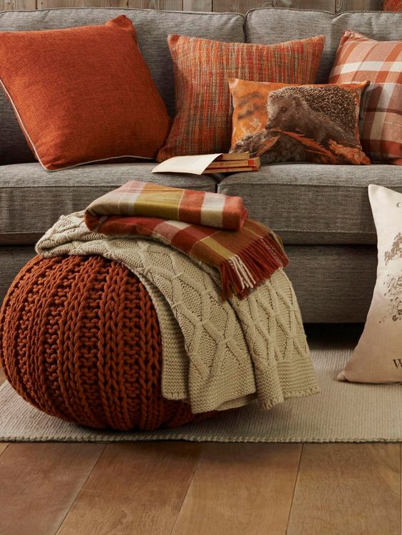Cozy living room with orange and plaid throw pillows, knit pouf with blankets, and a rustic decor on a grey couch.