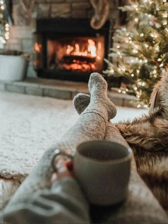 Cozy winter scene by a fireplace with a Christmas tree, person in warm socks holding coffee, holiday relaxation vibes.