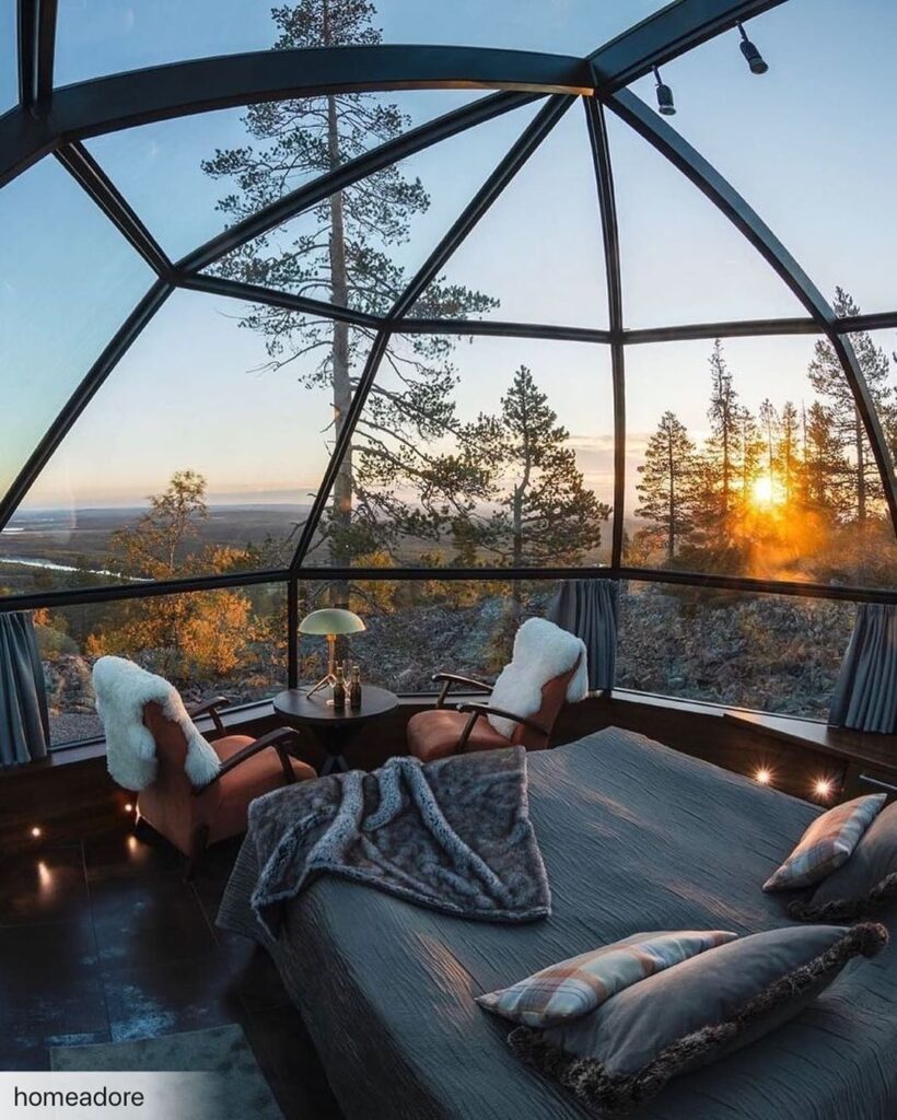 Cozy geodesic dome bedroom with panoramic forest view and sunset light, featuring plush chairs and soft blankets.