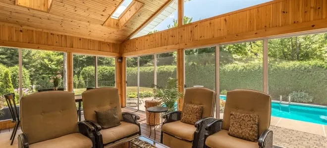 Cozy sunroom with wooden interior, comfortable seating, and large windows overlooking outdoor pool area and lush greenery.
