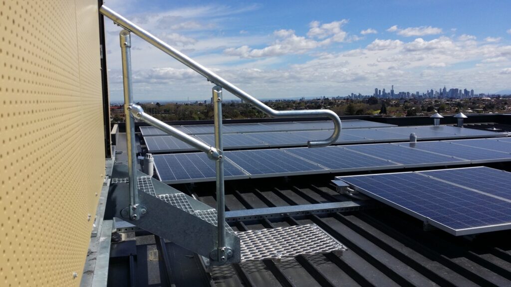 Rooftop solar panels under a blue sky with a distant skyline and safety railing in the foreground.