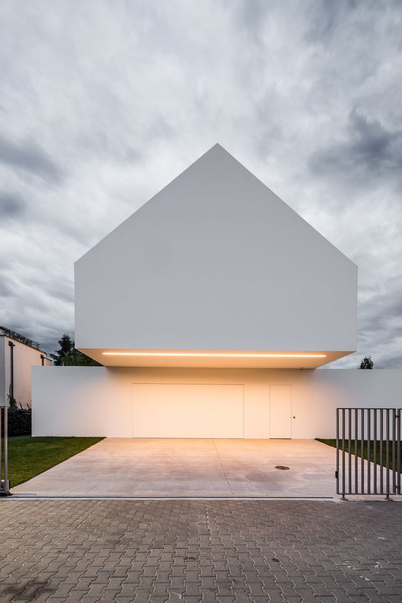 Modern minimalist house with a white geometric facade and integrated garage, set against an overcast sky.