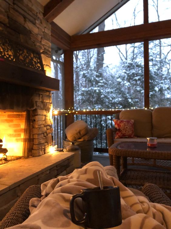Cozy winter scene with a lit fireplace, blankets, and hot drink on a porch with snowy trees outside. Comfort and warmth.