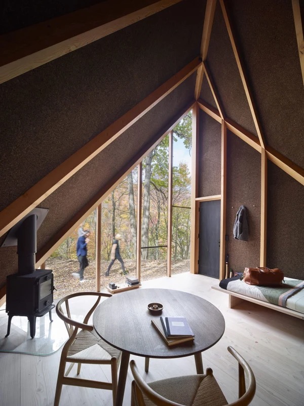 Cozy cabin interior with large windows, natural wood design, round table, and a forest view. Two people walking outside.